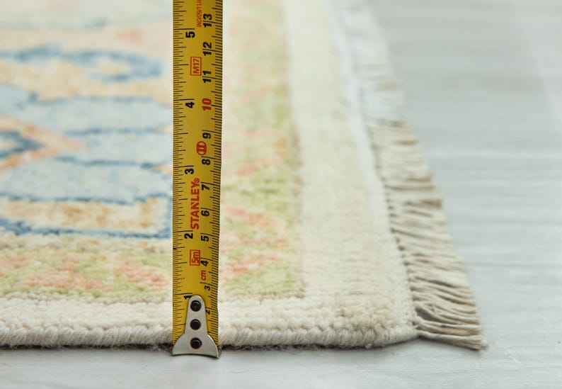 May include: Close-up of a section of a rug with a cream-colored border and fringe. The rug has a light blue and peach pattern. A yellow measuring tape shows the rug is approximately 13 inches long.