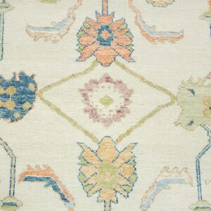 May include: Close-up of a cream-colored rug with an intricate floral pattern. The design features stylized flowers in shades of blue, orange, green, and pink, connected by a network of geometric lines. The rug has a textured surface.