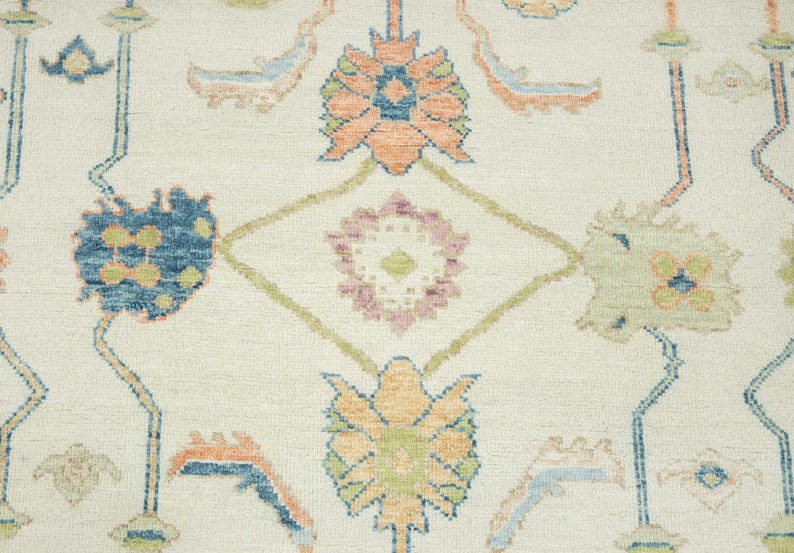 May include: Close-up of a cream-colored rug with an intricate floral pattern. The design features stylized flowers in shades of blue, orange, green, and pink, connected by a network of geometric lines. The rug has a textured surface.