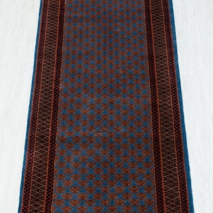 May include: A long, rectangular runner rug featuring a vibrant blue field decorated with a repeating pattern of dark rust-colored floral motifs. The rug is framed by a wide, dark border with intricate geometric designs in black and rust tones.