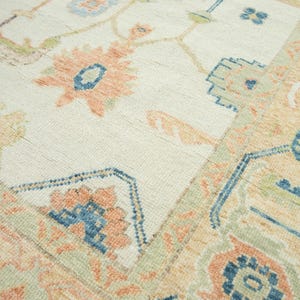 May include: Close-up of a cream-colored rug with a detailed floral pattern. The design features peach, blue, and green accents. The rug has a textured surface and a traditional, ornate style.
