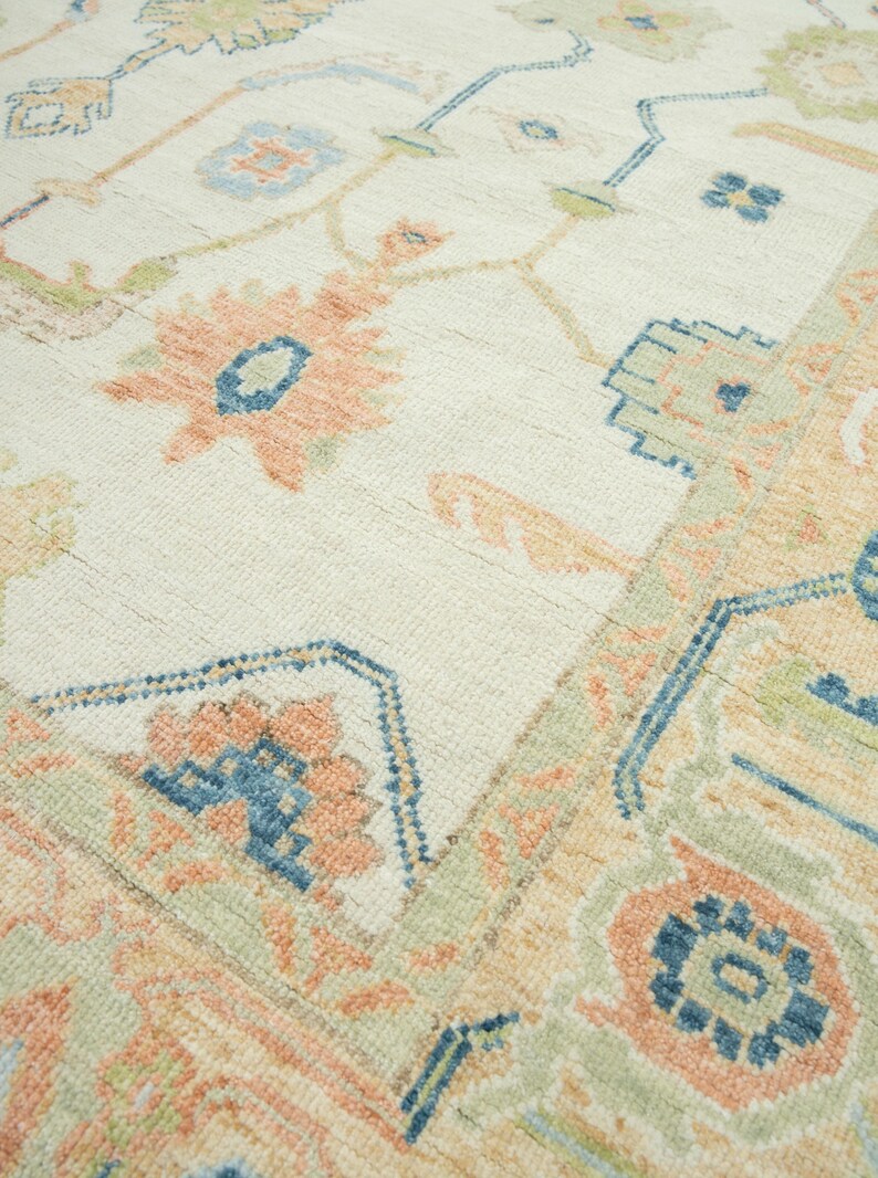 May include: Close-up of a cream-colored rug with a detailed floral pattern. The design features peach, blue, and green accents. The rug has a textured surface and a traditional, ornate style.