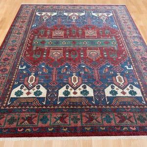 Puede incluir: Una alfombra rectangular que presenta un campo central de color rojo intenso con intrincados patrones geométricos en azul, verde y blanco. El borde muestra un motivo floral repetitivo en rojo y azul, acabado con flecos blancos.