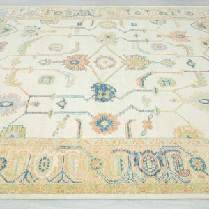 May include: Cream-colored area rug with a detailed floral pattern in shades of blue, orange, green, and pink. The rug has a beige border with geometric designs. A decorative floor covering for a living room or bedroom.