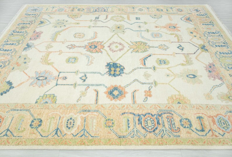 May include: Cream-colored area rug with a detailed floral pattern in shades of blue, orange, green, and pink. The rug has a beige border with geometric designs. A decorative floor covering for a living room or bedroom.