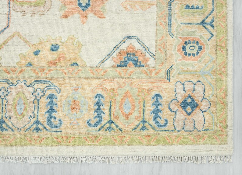 May include: Cream-colored area rug with a detailed floral pattern in shades of peach, blue, and green. The rug features a border with a fringe along the bottom edge. The design is intricate and traditional, suitable for home decor.