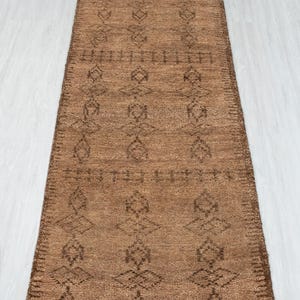 Puede incluir: Una alfombra pasillera larga y rectangular en tono marrón medio, con un patrón geométrico repetitivo de color marrón oscuro formado por rombos y líneas horizontales. La alfombra tiene una superficie texturizada y flecos blancos.