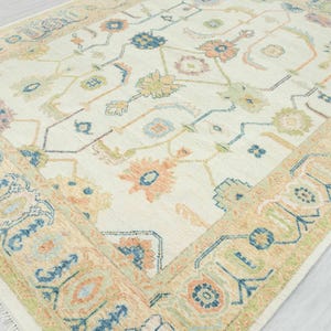 May include: Cream-colored area rug with a floral pattern in shades of peach, blue, green, and tan. The rug has a detailed border with geometric and floral designs. The rug's design is symmetrical and the edges are fringed.