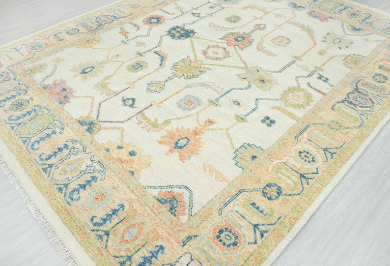 May include: Cream-colored area rug with a floral pattern in shades of peach, blue, green, and tan. The rug has a detailed border with geometric and floral designs. The rug's design is symmetrical and the edges are fringed.