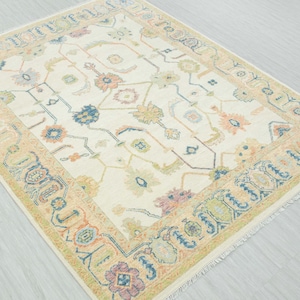 May include: Cream-colored area rug with a detailed floral pattern in shades of blue, orange, green, and purple. The rug has a border with a repeating design and fringe along the edges. A decorative floor covering.