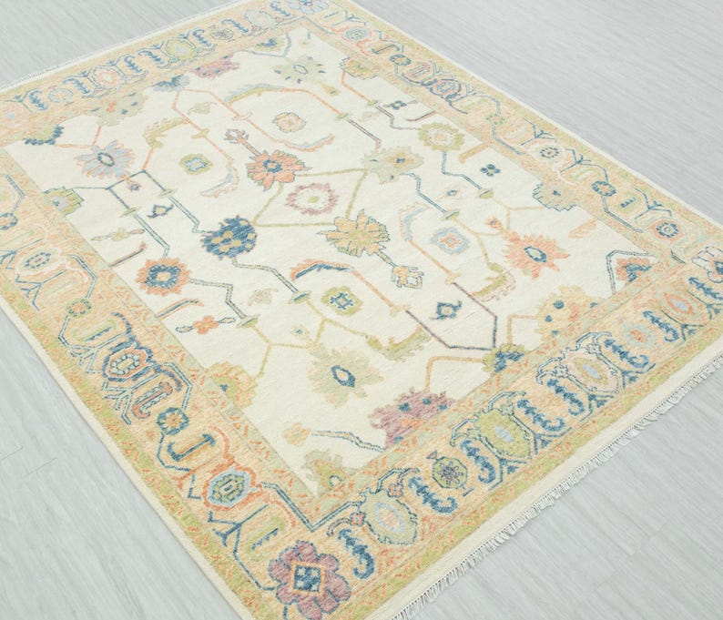 May include: Cream-colored area rug with a detailed floral pattern in shades of blue, orange, green, and purple. The rug has a border with a repeating design and fringe along the edges. A decorative floor covering.