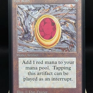 Può includere: Una carta Mox Ruby con un ciondolo dorato con una grande gemma rossa su uno sfondo marmorizzato. La carta recita "Mono Artifact" e include testo sull'aggiunta di mana rosso. Il nome dell'artista, Dan Frazier, è anch'esso visibile.