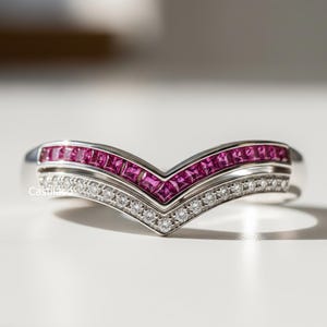 Peut inclure: Une bague en argent en forme de V avec une rangée de pierres roses taillées en carré et une rangée de petites pierres claires et rondes. La bague est posée sur une surface blanche.