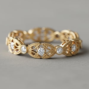 以下が含まれることがあります： 精巧なフィリグリーデザインのゴールドリング。リングは、それぞれ小さな丸い透明な宝石で飾られた、一連の葉の形をした要素を特徴としています。リングはニュートラルなグレーの背景に配置されています。