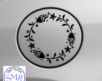 May include: A black decal featuring a circular design with a nautical theme. The design includes jellyfish, starfish, and leafy vines arranged in a wreath pattern. The decal is on a silver surface.
