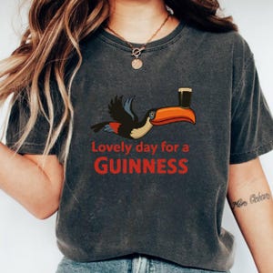 May include: Dark grey t-shirt with a cartoon toucan carrying a Guinness pint. The red text reads "Lovely day for a GUINNESS". A casual wear item.