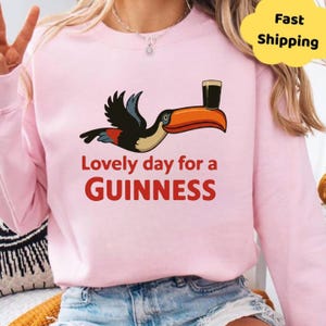 May include: Pale pink sweatshirt featuring a cartoon toucan with a pint of Guinness. The text "Lovely day for a Guinness" is printed in red. A yellow cloud with "Fast Shipping" is in the upper right.
