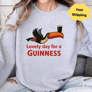 May include: A light grey sweatshirt with a toucan design carrying a pint of Guinness. The text "Lovely day for a Guinness" is in red. The toucan has a large orange beak and is in flight.