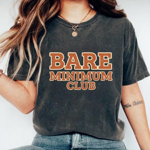 May include: Dark gray t-shirt with the words "BARE MINIMUM CLUB" in a brown and cream-colored font. The shirt has a vintage, worn look. The image shows the upper body of a person wearing the shirt.