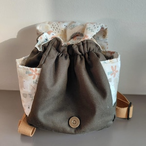 Puede incluir: Una mochila de cordón con un panel central de tela verde oliva oscuro sólido y un cierre de broche metálico. Los laterales y la solapa superior están hechos de tela color crema con un patrón repetitivo de pequeñas flores y hojas naranjas.