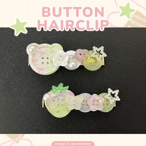 Może przedstawiać: Dwie ozdobne spinki do włosów z guzikami. Jedna spinka ma wzór misia, druga truskawki. Obie mają perłowe akcenty, detale guzików i napis "BUTTON HAIRCLIP". Spinki są w pastelowych kolorach.