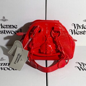 May include: A bright red, glossy handbag with a unique design. The bag features a structured shape with a top handle and a shoulder strap. A tag with the brand name is attached. The bag is set against a white background.