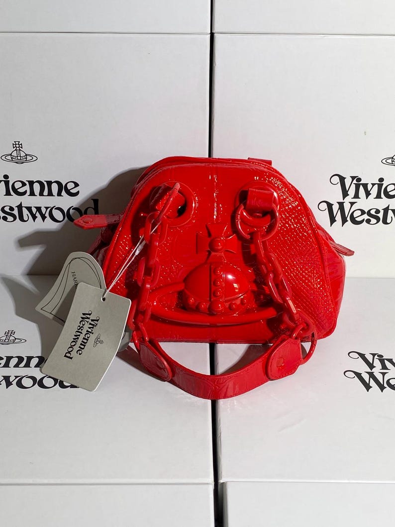 May include: A bright red, glossy handbag with a unique design. The bag features a structured shape with a top handle and a shoulder strap. A tag with the brand name is attached. The bag is set against a white background.