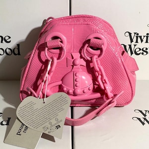 May include: A bright pink, small backpack with a textured, crocodile-like pattern. The bag features a prominent orb emblem and chain straps. A tag with text hangs from the bag. The bag is sitting on top of a white box with the words "Vivienne Westwood" printed on it.