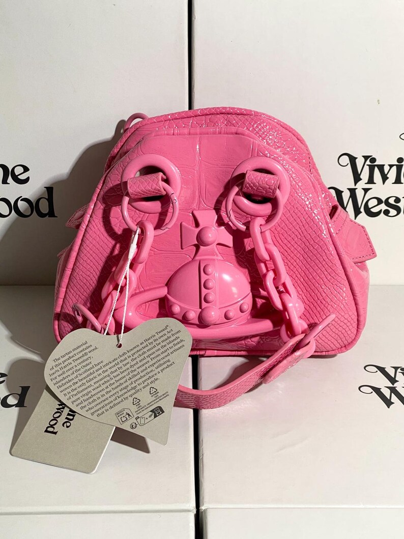May include: A bright pink, small backpack with a textured, crocodile-like pattern. The bag features a prominent orb emblem and chain straps. A tag with text hangs from the bag. The bag is sitting on top of a white box with the words "Vivienne Westwood" printed on it.