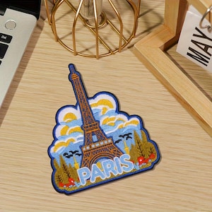 May include: Embroidered patch featuring the Eiffel Tower in Paris, France. The patch has a blue border and depicts the tower in brown and gold, with blue sky and clouds. The word "PARIS" is written in white letters at the bottom.
