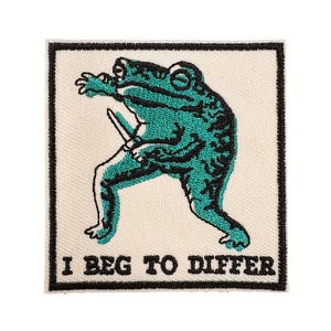 May include: Embroidered patch featuring a teal frog holding a knife, with the phrase "I BEG TO DIFFER" in black text. The frog has black detailing and is set against an off-white background with a black border. Ideal for jackets, bags, or other fabric items.