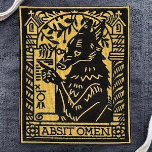 May include: Black and gold embroidered patch featuring a wolf-like creature with glasses, pointing. The patch has the text "ABSIT OMEN" at the bottom and is surrounded by ornate, gothic-style designs. The patch is rectangular.
