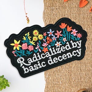 May include: Black embroidered patch with a floral design and the text "Radicalized by basic decency" in white. The flowers are in shades of yellow, pink, orange, and blue, with green leaves. The patch has a scalloped edge.