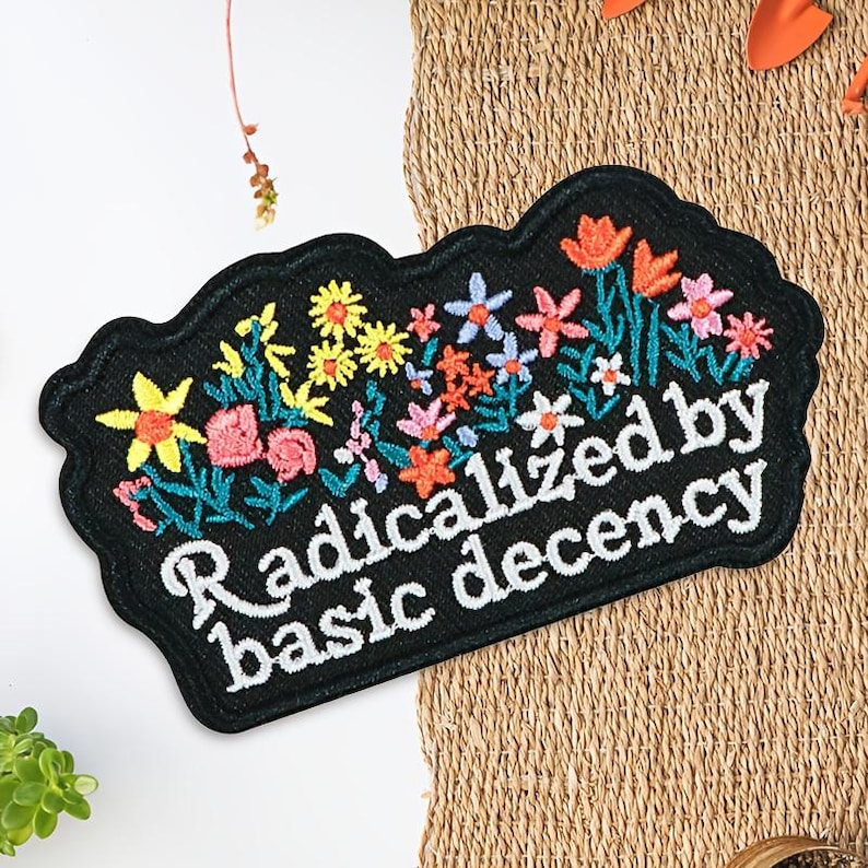 以下が含まれることがあります： 黒色の刺繍パッチ。花柄のデザインと「Radicalized by basic decency」の白文字。花は黄色、ピンク、オレンジ、青色で、緑の葉があります。パッチはスカラップエッジです。