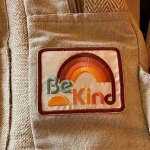 May include: A rectangular patch with a rainbow and the words "Be Kind" embroidered in various colors. The patch is bordered in a dark red and is sewn onto a beige fabric.