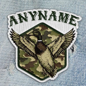 May include: Embroidered patch featuring a duck with outstretched wings, set against a camouflage background within a green-bordered hexagon. The text "ANYNAME" arches above the duck. The patch is stitched with white thread.
