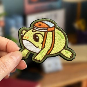 May include: Embroidered patch featuring a cartoon frog in shades of green, yellow, and brown. The frog is wearing a hat with orange and blue accents. The patch has a dark green border and is held by a hand.