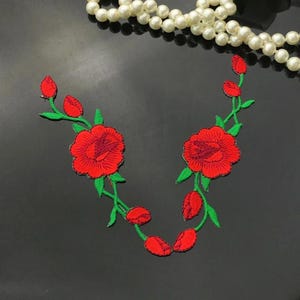 May include: Embroidered red rose patches with green stems and leaves. The patches feature detailed stitching and are arranged in a curved design. A strand of white pearls is in the background.