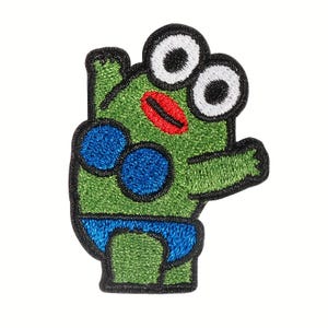 May include: Embroidered patch featuring a cartoon frog with large white eyes, a red mouth, and blue accents. The frog is green with a black outline. The patch is designed to be ironed on or sewn onto fabric.