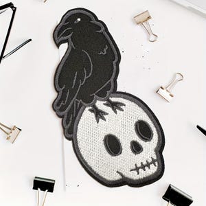 May include: Embroidered patch featuring a black raven perched atop a white skull. The raven and skull are outlined in gray. The patch is likely for clothing or accessories, with a gothic or Halloween theme.