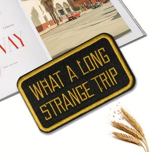 May include: Black rectangular patch with gold embroidered text that reads "WHAT A LONG STRANGE TRIP". The patch has a gold border and is set against a white background with a magazine and wheat stalks.