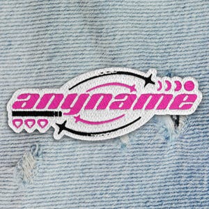 May include: Embroidered patch with the word "anyname" in pink, inside a white oval with black accents. The patch features stars, arrows, and other decorative elements. The patch is on a light blue denim background.