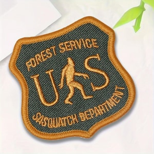 May include: Embroidered patch with a shield shape, featuring the text "FOREST SERVICE" and "SASQUATCH DEPARTMENT". The central design includes the letters "U S" and a Sasquatch figure, all in gold thread on a dark green background, with a gold border.