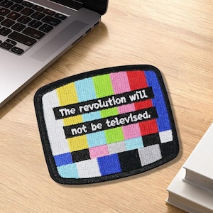 May include: Embroidered patch with a colorful test pattern design and the text "The revolution will not be televised." The patch has a black border and is rectangular with rounded corners. The background is a wooden surface.