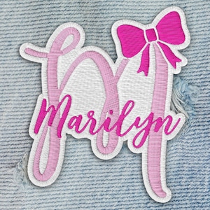 May include: Embroidered patch featuring the name "Marilyn" in a cursive font, with a pink bow accent. The patch has a white background and a pink outline, set against a denim background.