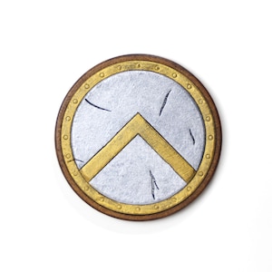 Può includere: Un emblema rotondo a forma di scudo, con un centro argentato e un gallone dorato. L'emblema ha un bordo marrone con rivetti dorati. Lo scudo ha un aspetto invecchiato con segni di graffio neri.