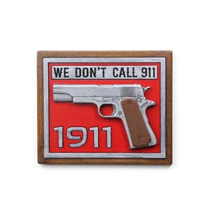 May include: A rectangular wooden sign with a red background and a silver-colored handgun. The text "WE DON'T CALL 911" is at the top, and "1911" is at the bottom. The sign has a brown wooden frame.