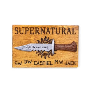 以下が含まれることがあります： 上部に「SUPERNATURAL」の文字が彫られた木製のプラーク。茶色の柄が付いた銀色のナイフが中央にあり、「SW DW CASTIEL MW JACK」という言葉の上にあります。背景は明るい茶色です。