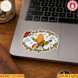 May include: An oval sticker featuring a person with long blonde hair playing an acoustic guitar. The text reads Love has ways, Bringing things back to life. A red cowboy hat, blue clouds, and musical notes surround the figure, all framed by a rope border.