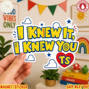 May include: A die-cut sticker featuring the yellow text I KNEW IT, I KNEW YOU TS with a blue outline. The design includes small blue stars, white cloud outlines, and a red heart shape containing the letters TS, all on a white background.